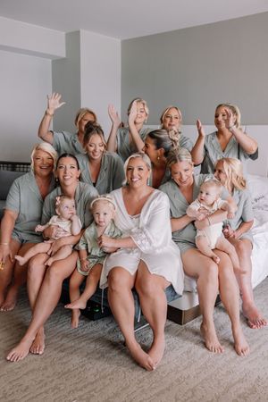 Bride in white robe poses with bridesmaids in sage green robes and babies on a bed, all smiling and waving.