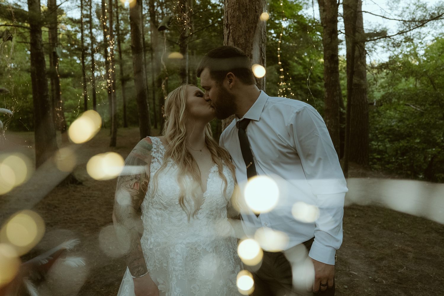 A bride and groom share a romantic kiss in a forest wedding setting with bokeh fairy lights in the foreground.