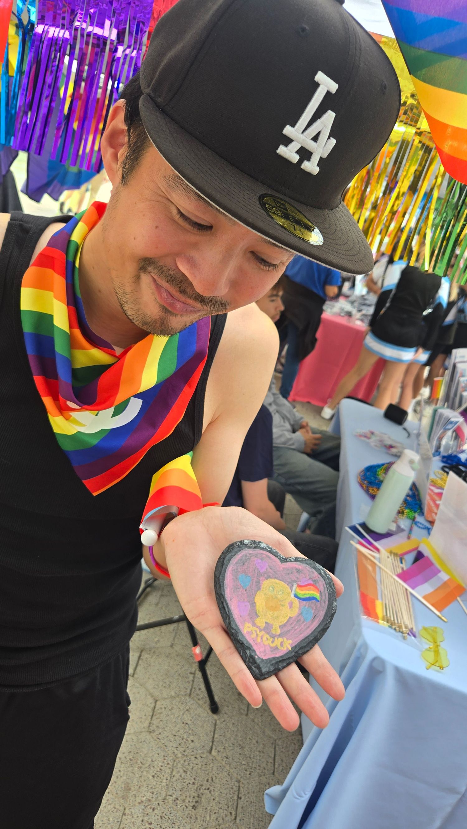 Man in LA cap holding a heart-shaped Pride rock at an outdoor LGBTQ+ Pride event with rainbow decorations.
