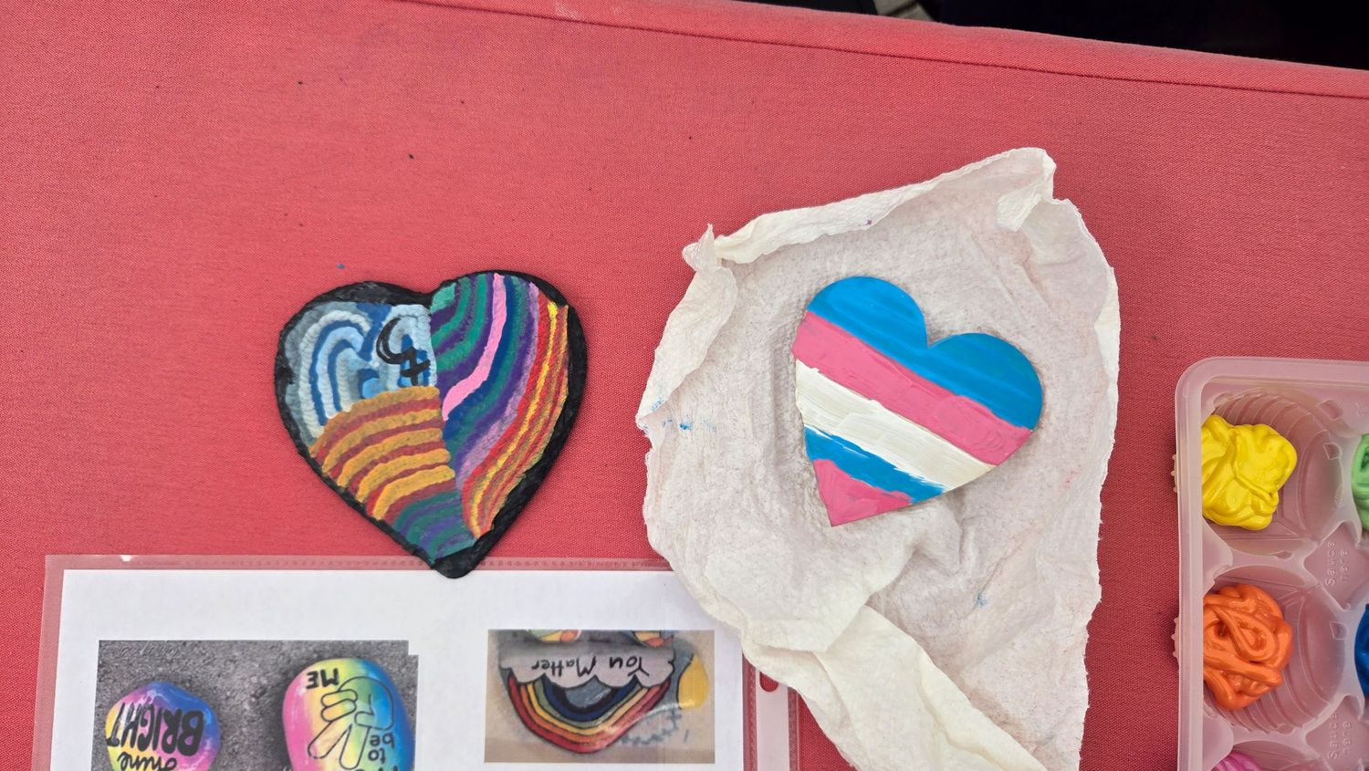Heart-shaped patches featuring rainbow and transgender pride flag designs displayed on a pink surface with paint palette.