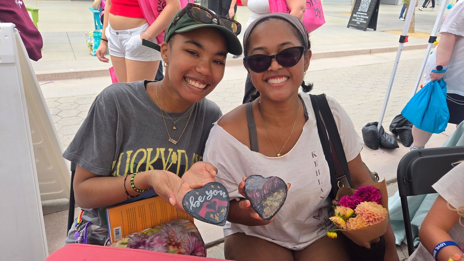 Two smiling women posing together outdoors at a festival, holding colorful handmade crafts and gifts.