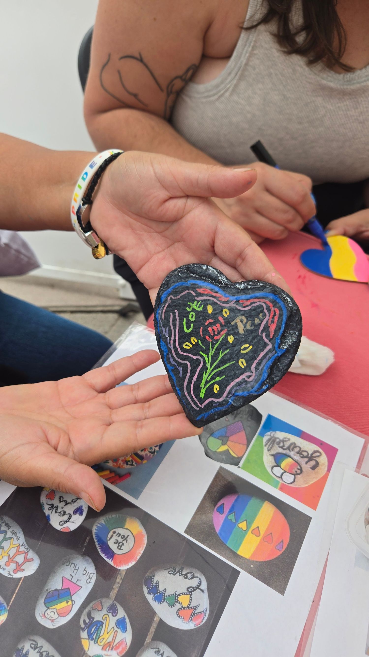 Hands holding a heart-shaped rock painted with colorful flowers on black background at a rock painting craft event.