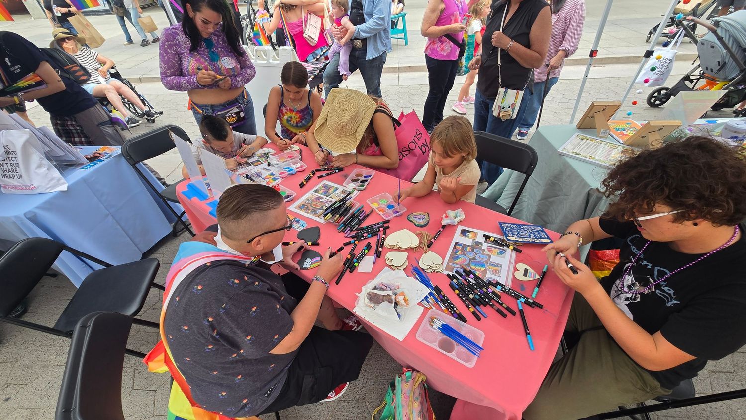 Children and adults engage in a craft activity at a pink table during an outdoor community event.