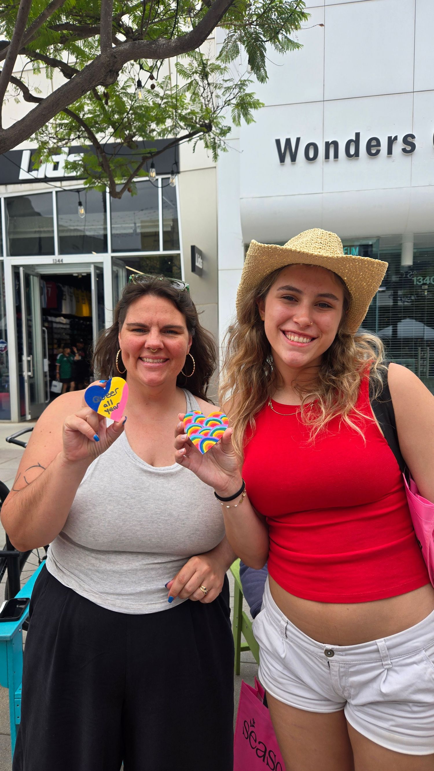 Two smiling women pose outdoors holding colorful macarons near a Wonders store, one wearing a straw hat and red crop top.