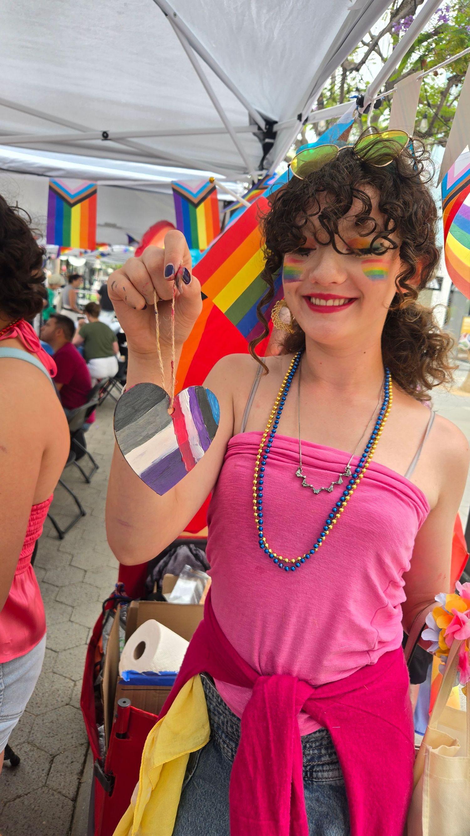 Person at Pride event in pink top holding asexual flag heart necklace, with rainbow face paint and colorful beaded necklaces.