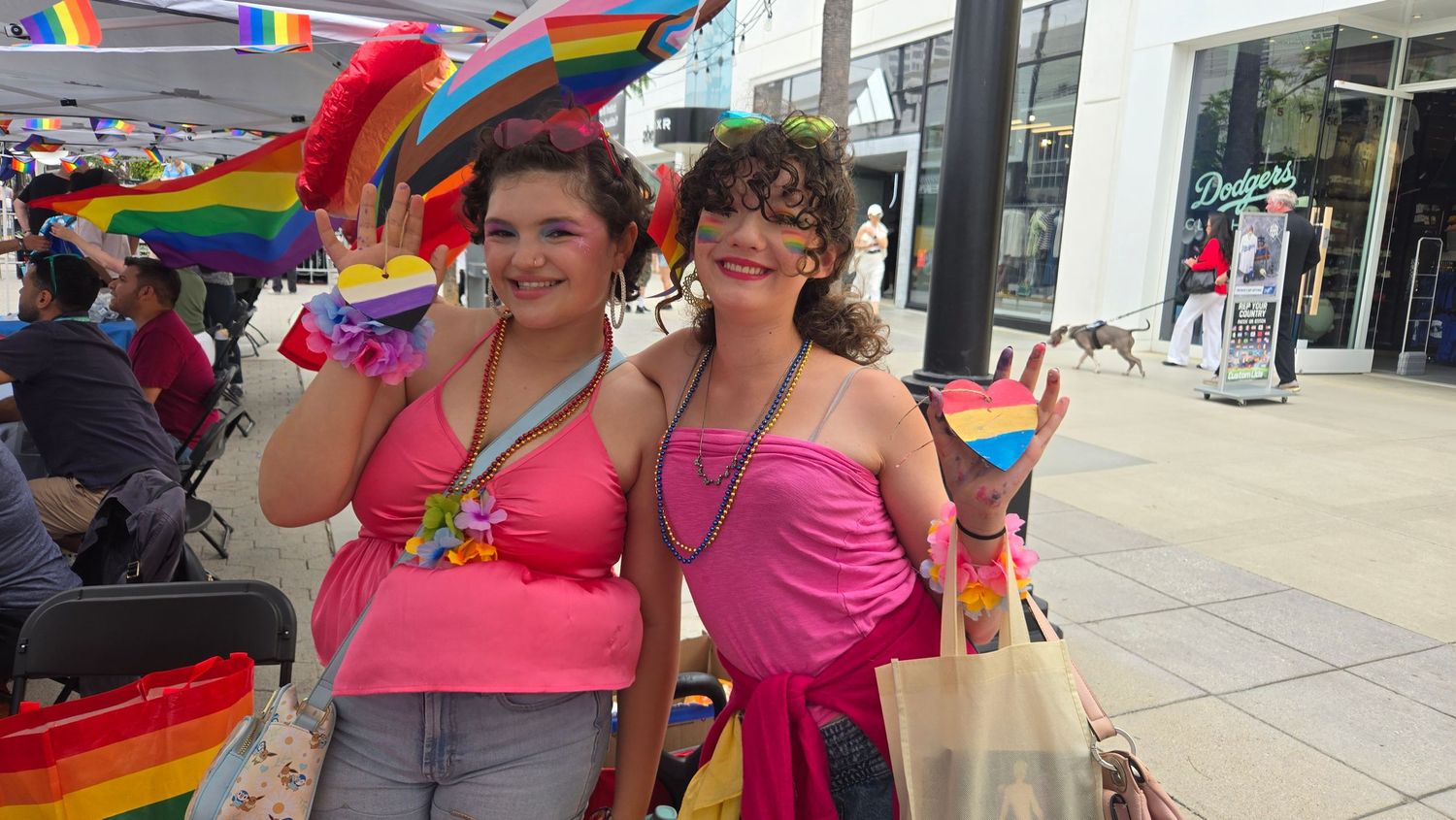 Two women in pink outfits smile at an outdoor Pride event surrounded by rainbow flags and decorations.