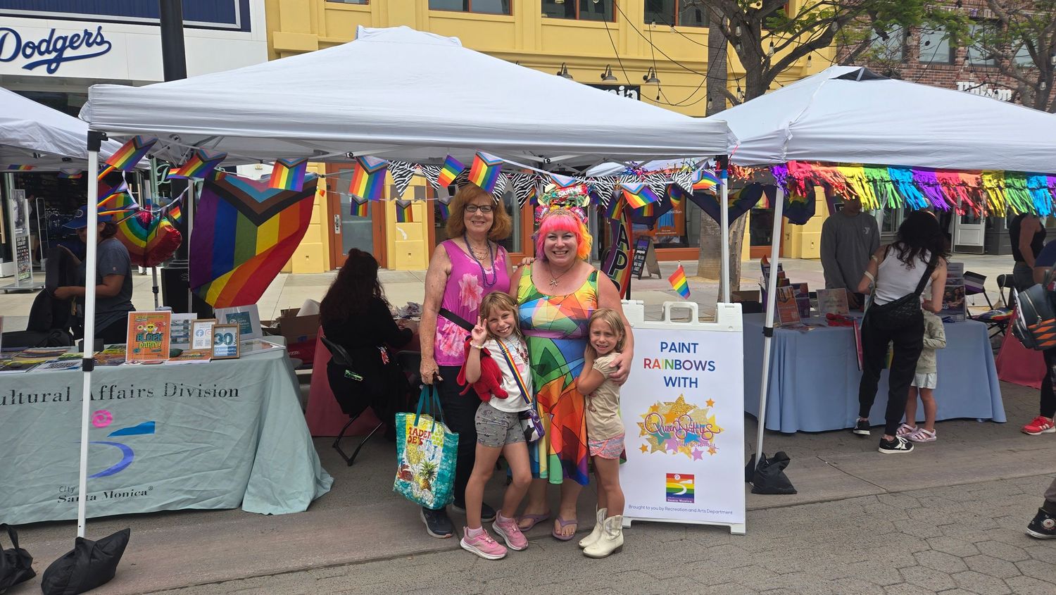 Two women and children pose at a colorful Pride festival booth decorated with rainbow flags and banners outdoors.