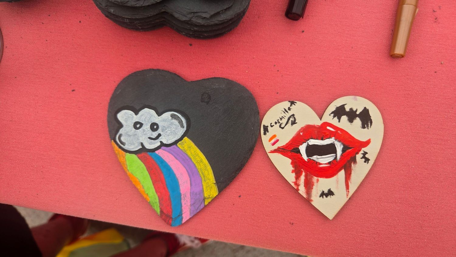 Two hand-painted heart-shaped crafts on a pink surface, one with a rainbow cloud and one with vampire lips and a bat.