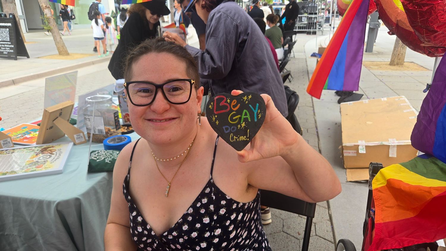 Woman with glasses holding a painted rock at a Pride event with rainbow flags in the background.