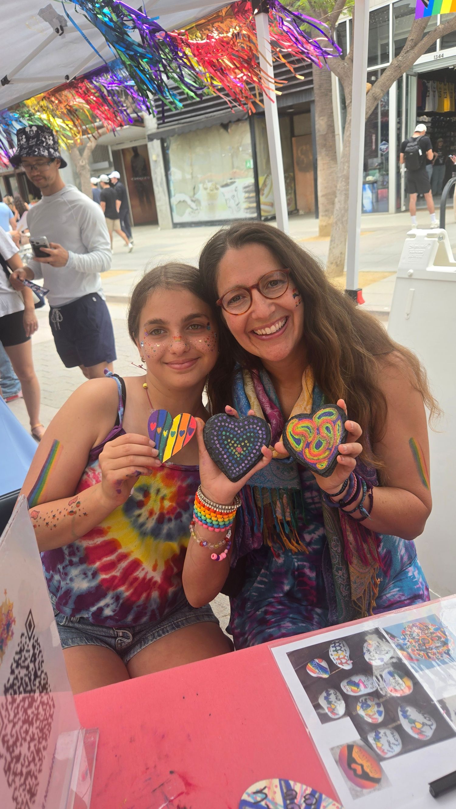 Two smiling women hold colorful painted rocks at a Pride festival booth, wearing tie-dye outfits with rainbow decorations overhead.