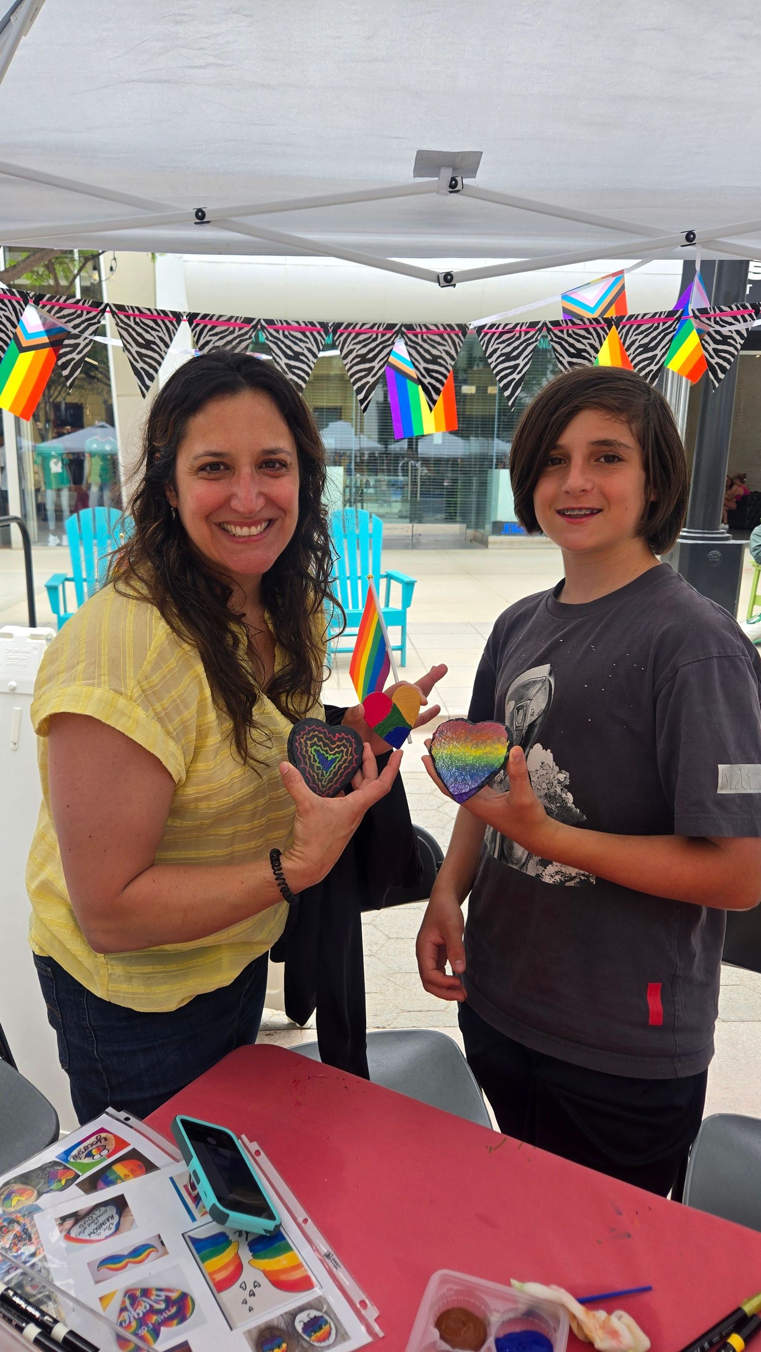 Woman and child proudly display rainbow heart crafts at a colorful Pride-themed art booth with festive decorations.
