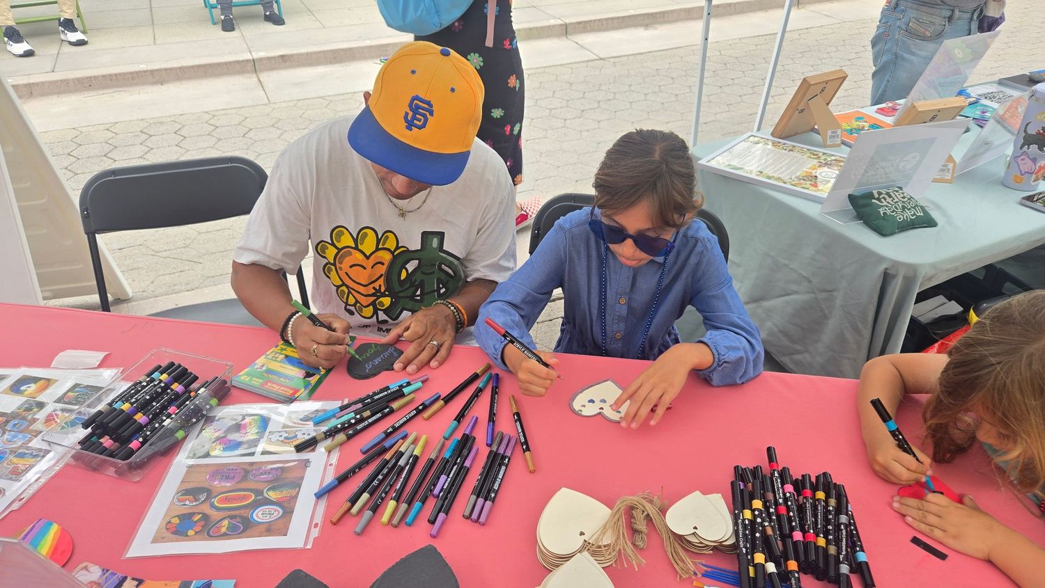 Children and adult doing arts and crafts at a table with markers and paint at an outdoor community event.