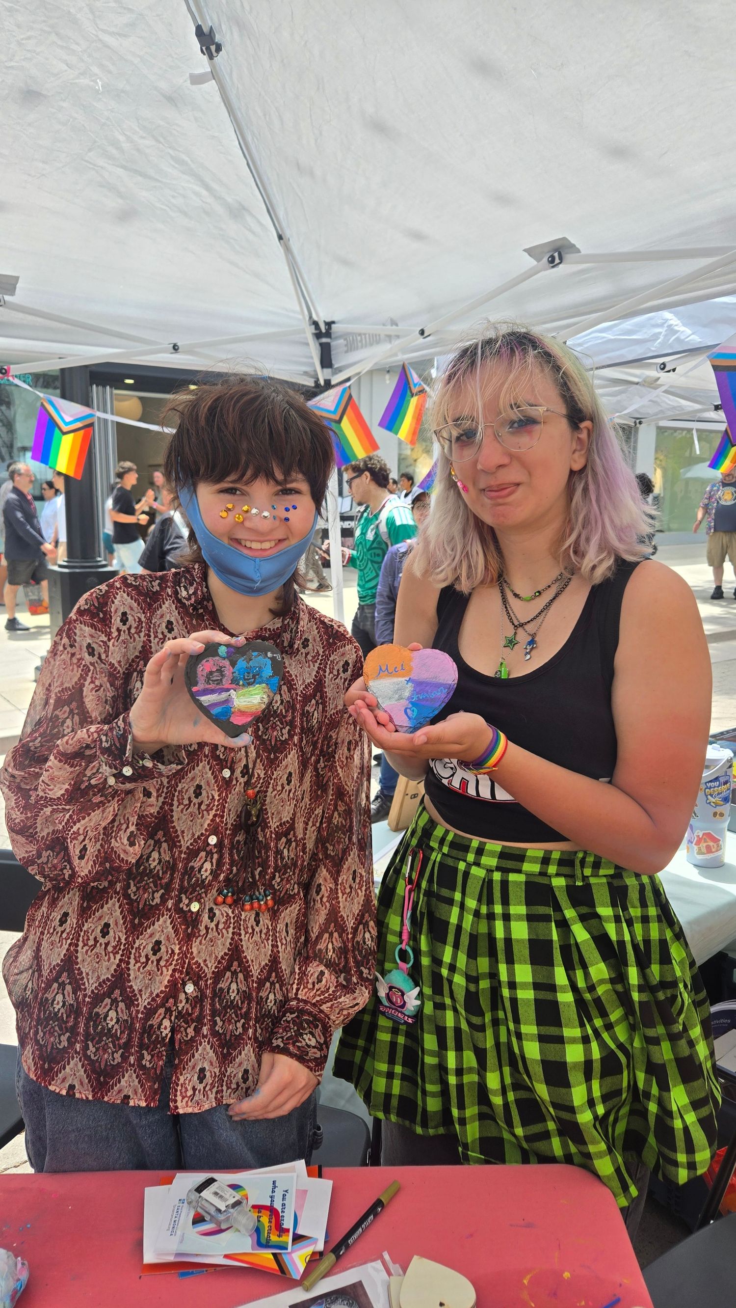 Two people holding painted heart rocks at a Pride event, with rainbow decorations and a tent in the background.