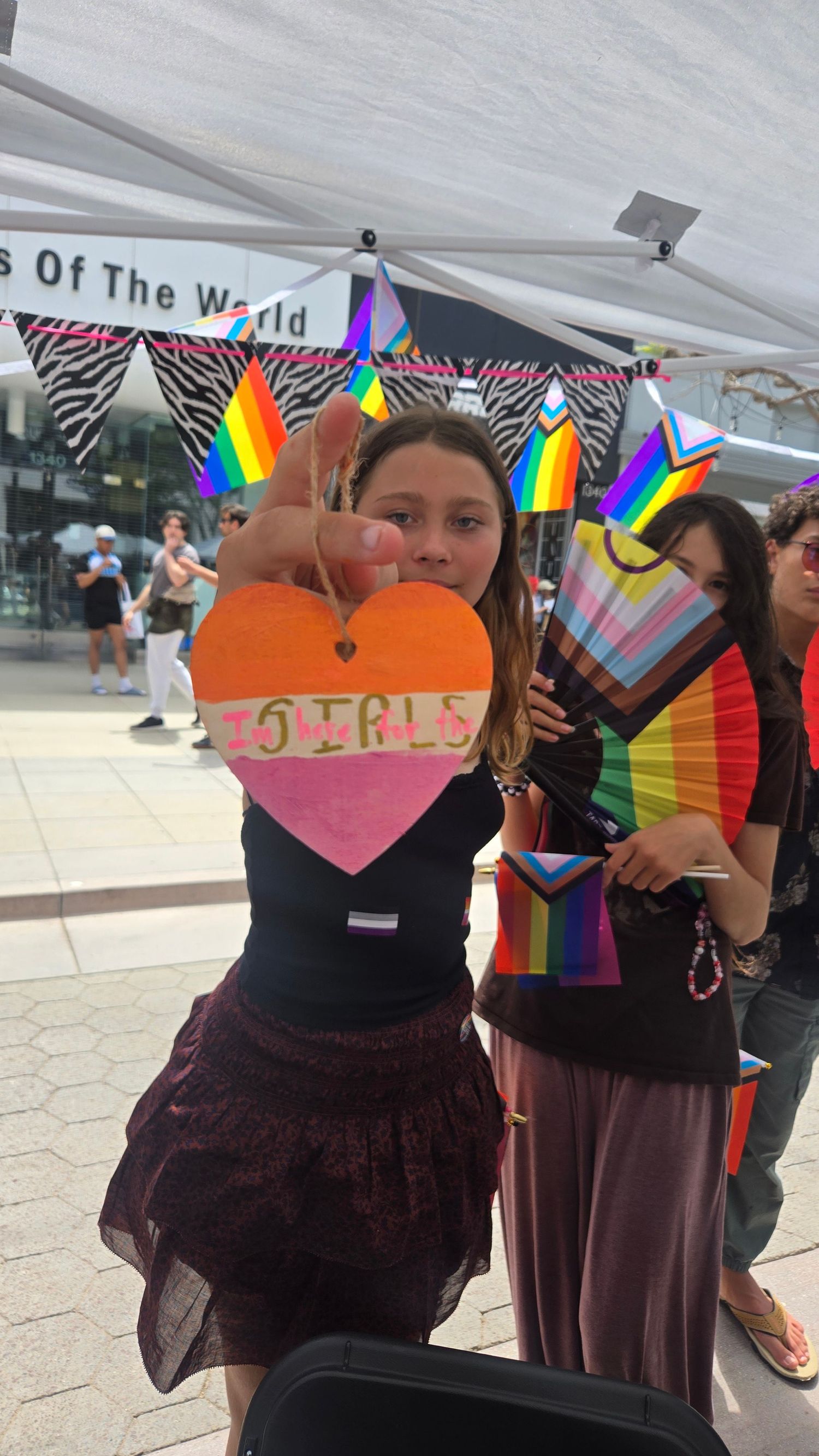 Young woman holds a heart-shaped sign reading 'Girls' at a Pride event with rainbow flags and decorations.