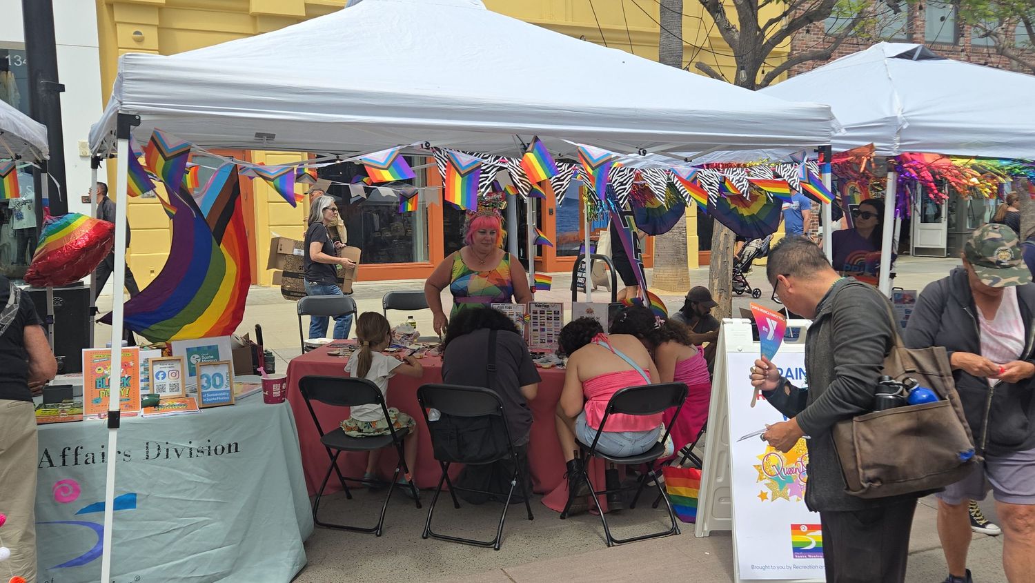 Colorful Pride festival booth decorated with rainbow flags and bunting, with attendees gathered under a white tent outdoors.