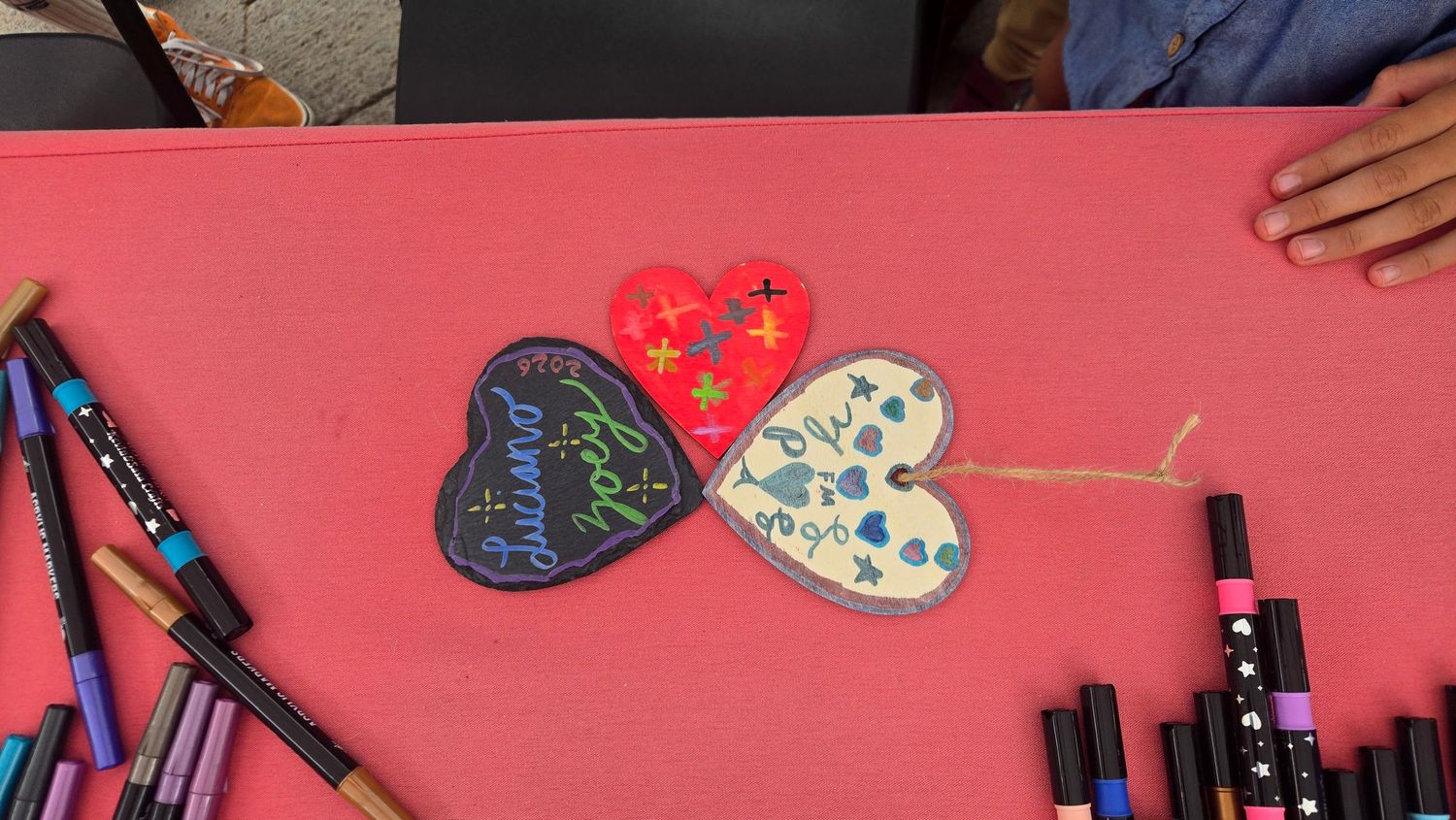 Three decorated heart-shaped ornaments connected by twine on a pink surface, surrounded by colorful markers.