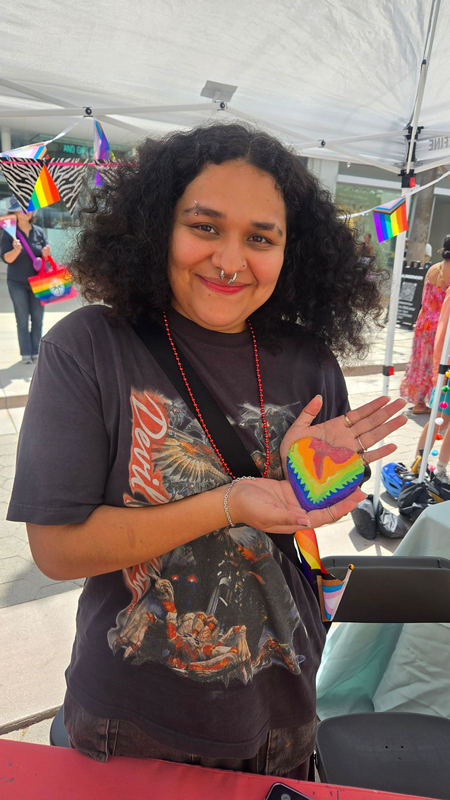 Person holding a rainbow heart-shaped painted rock at a Pride event outdoor vendor booth.
