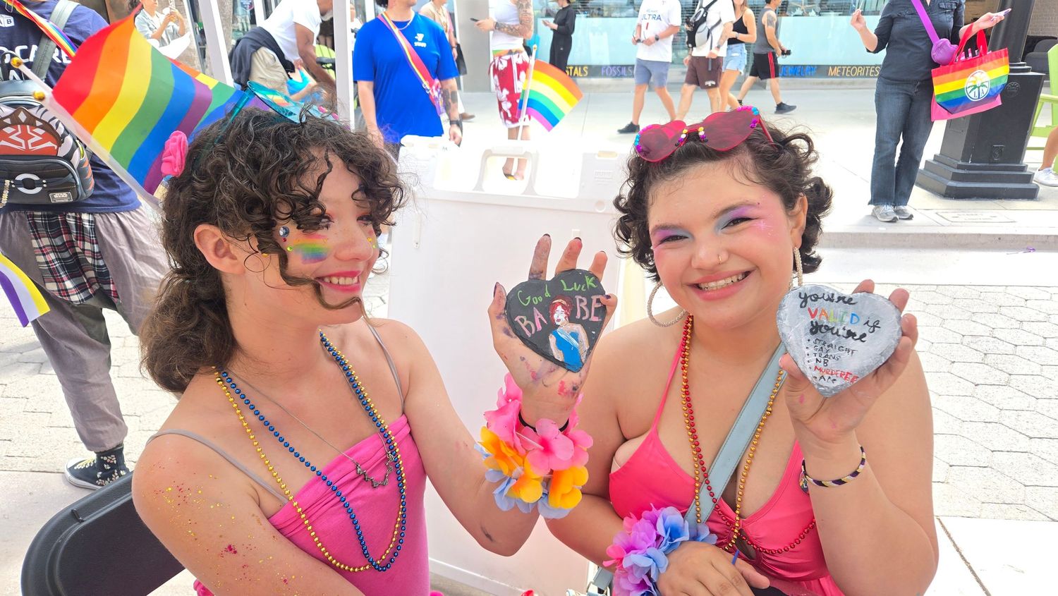 Two women in pink bikini tops holding painted rocks at a Pride parade with rainbow flags.