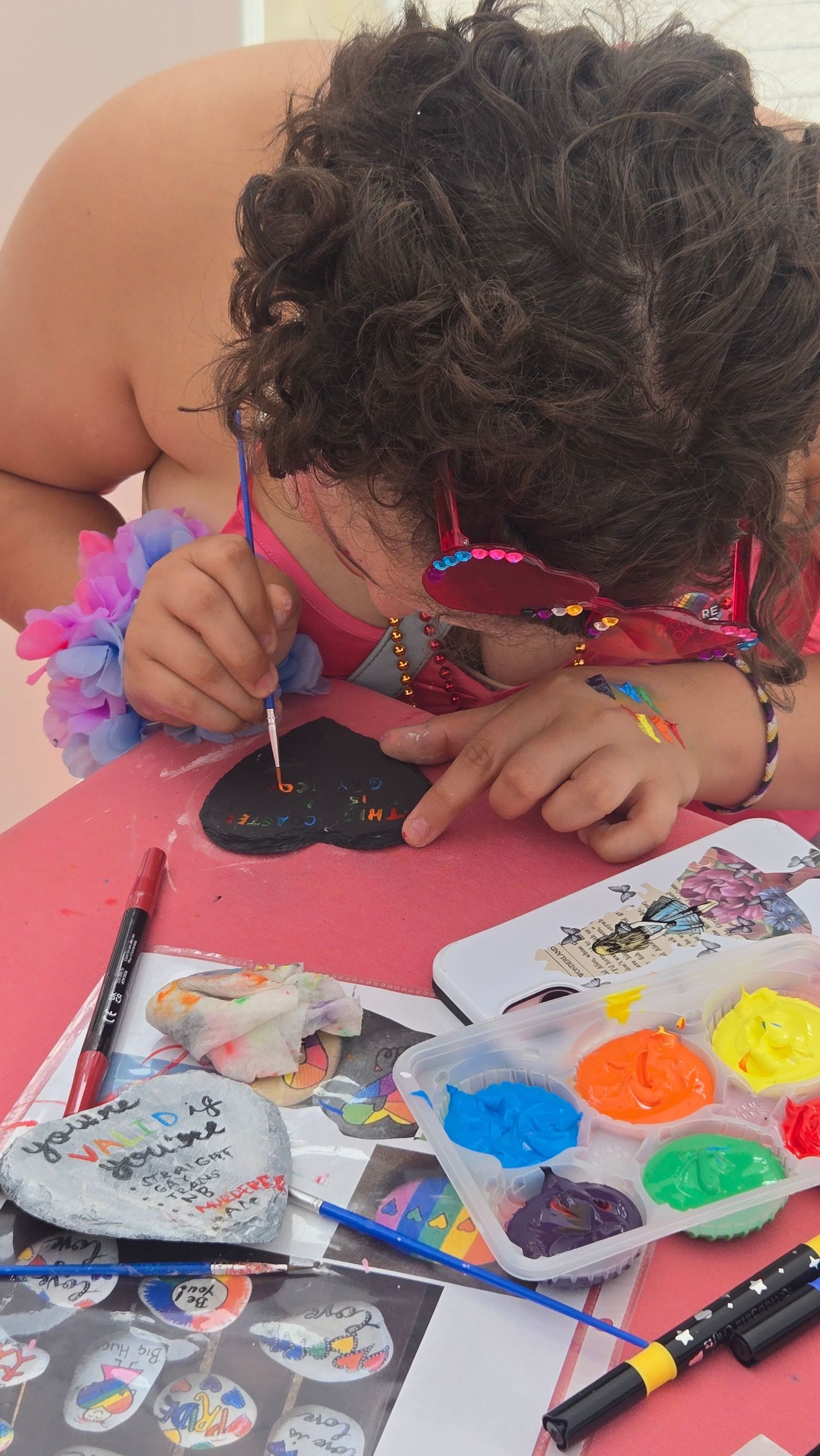Child painting a black rock with colorful paints at a craft table during an outdoor art activity.