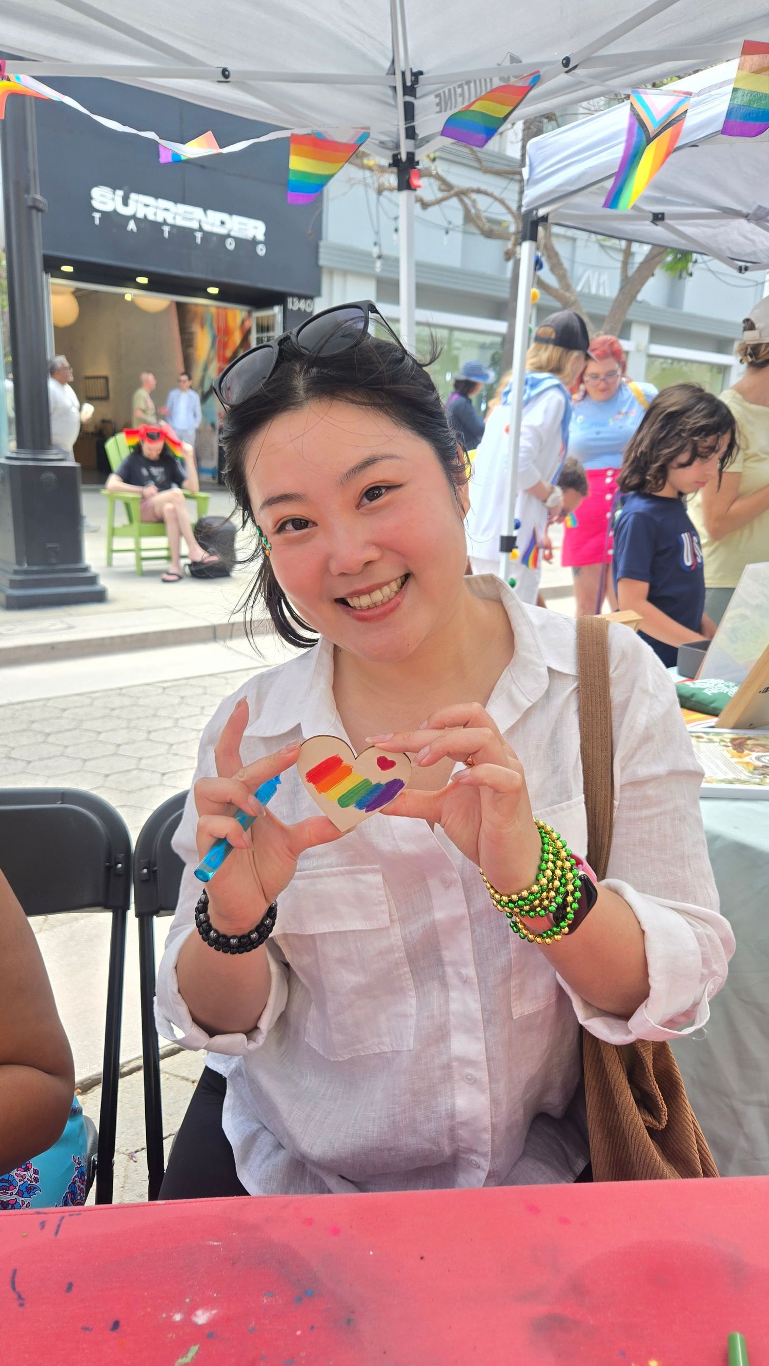 Smiling woman holding a rainbow-painted rock at an outdoor Pride event with flags in background.