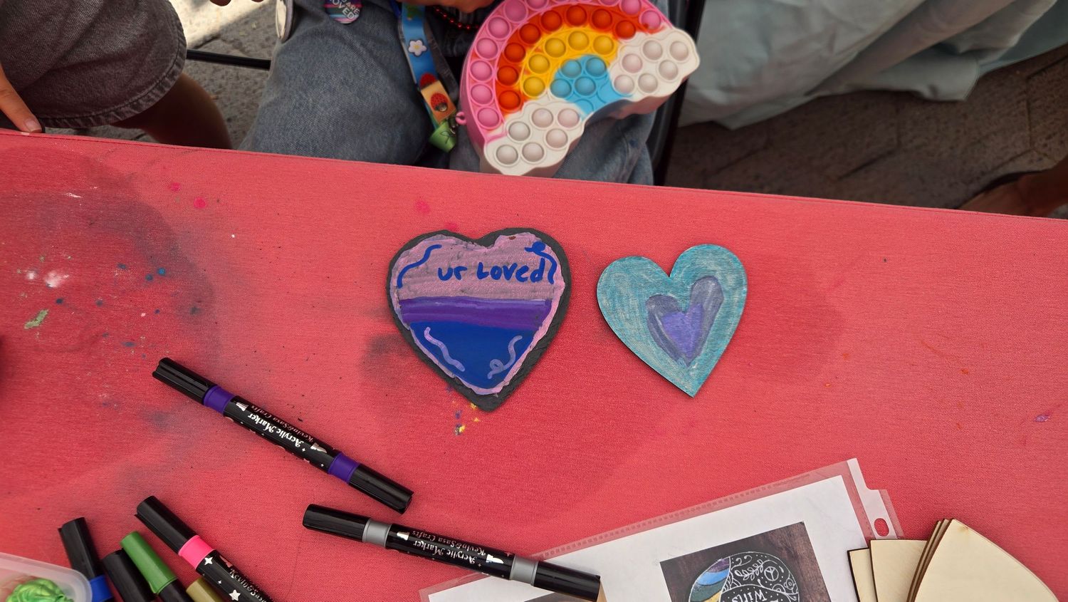 Handmade clay heart pins with colorful marker designs on a red table, one reading ur loved, surrounded by art supplies.