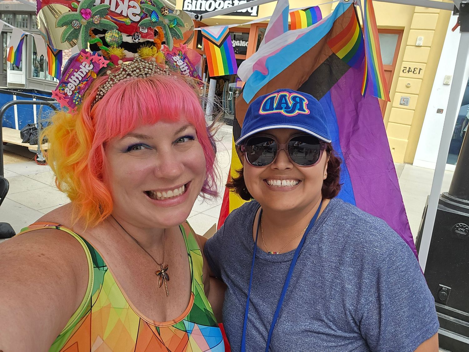 Two women smiling at Pride event, one in pink wig and rainbow outfit, one in blue cap, with Pride flags backdrop.