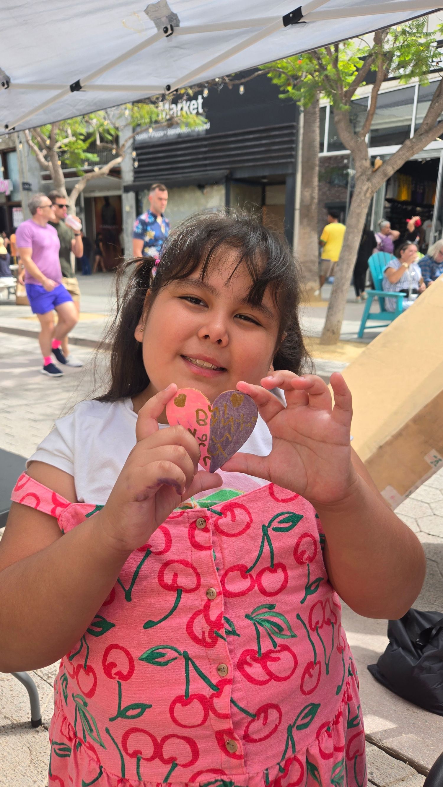 A young girl in a cherry-print dress proudly holds a heart-shaped craft at an outdoor market event.