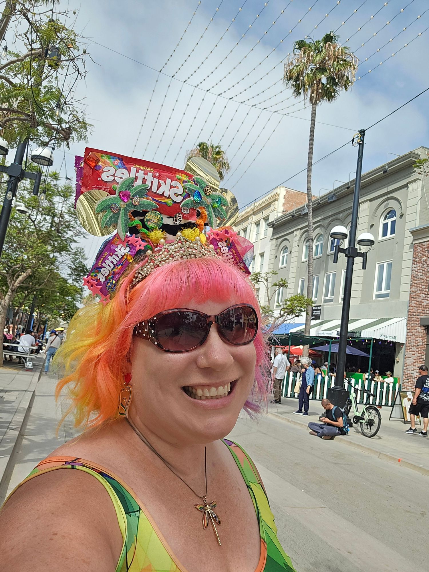 Woman with pink-orange hair and Skittles candy headpiece smiles outdoors on a sunny street with palm trees.