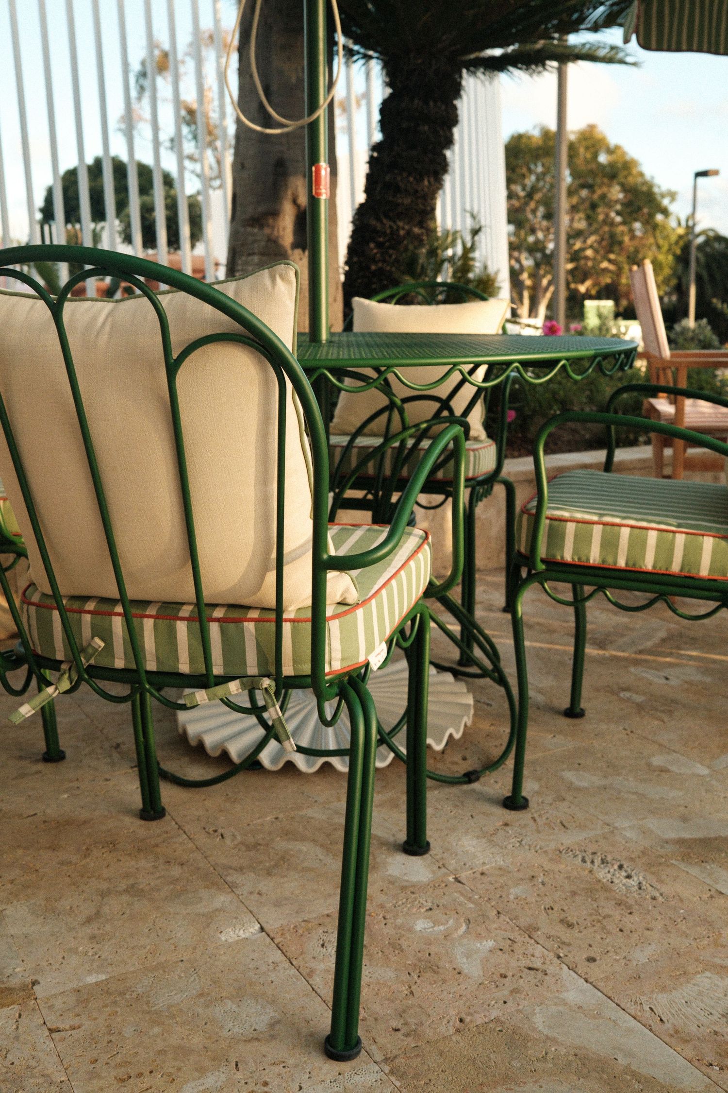 Green wrought iron patio chairs with striped cushions arranged around a table on a stone terrace near palm trees.