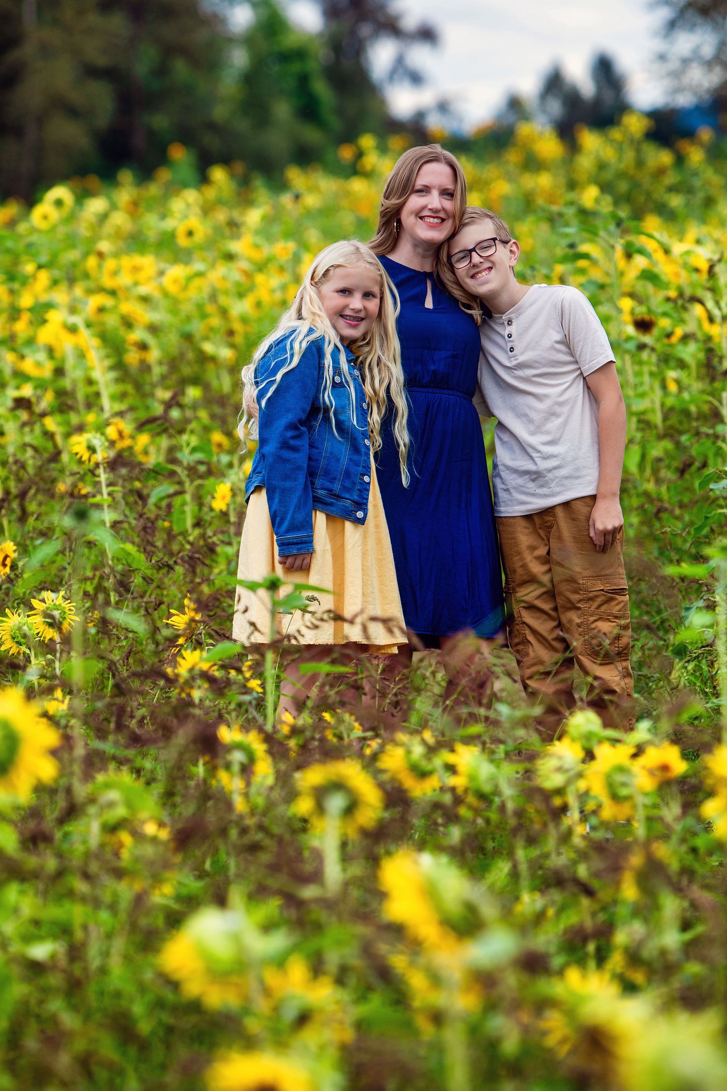 A mother and two children smile together in a vibrant sunflower field, the girl in a denim jacket and the boy in glasses.