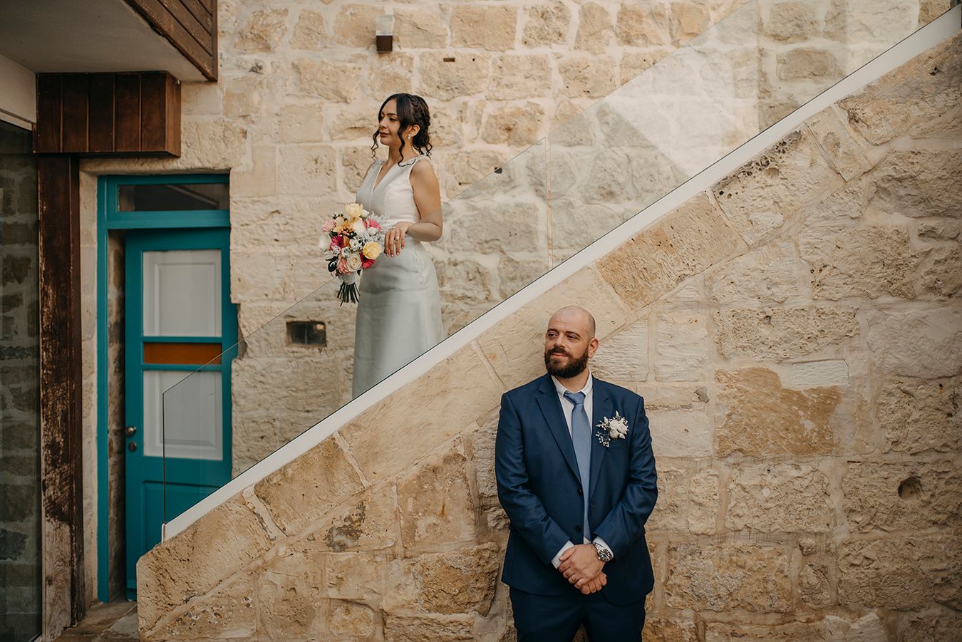 Bride stands on stone staircase holding bouquet while groom waits below in navy suit at outdoor wedding venue.