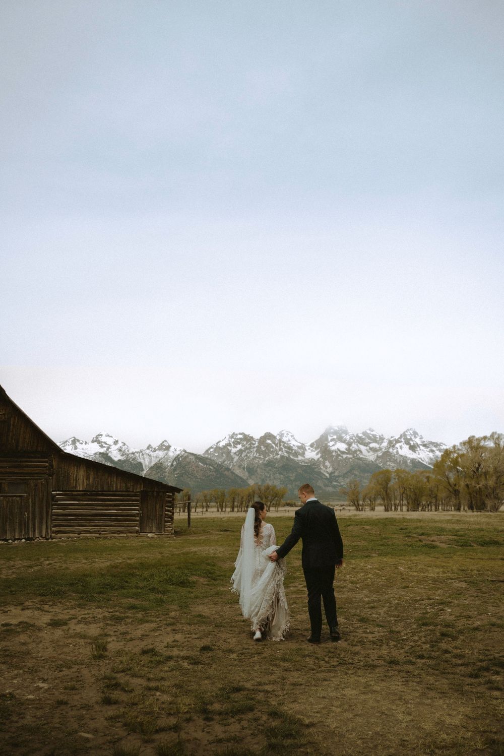 Bride and groom hold hands walking toward snow-capped Grand Teton mountains near rustic barn.