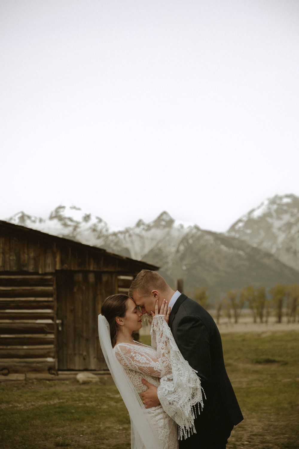 Bride and groom share a romantic kiss with snow-capped Grand Teton mountains and rustic barn backdrop.