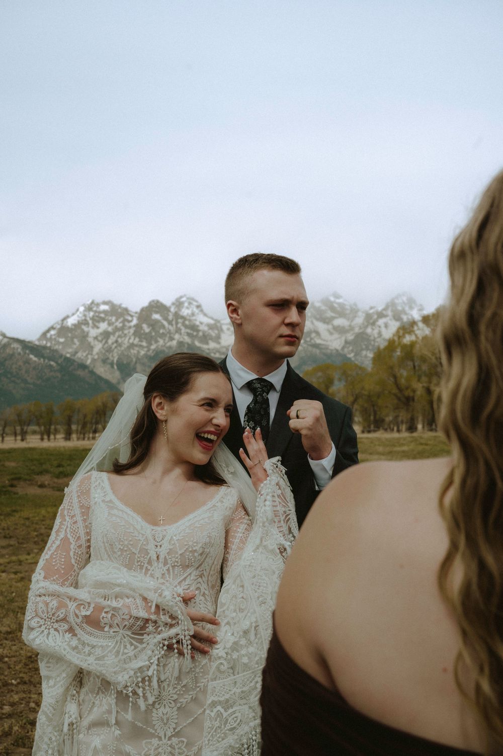 Bride in lace gown laughing with groom in dark suit at outdoor wedding with Grand Teton mountains backdrop.