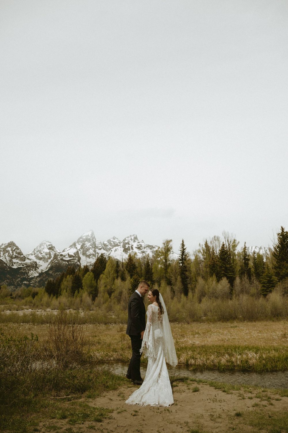 Bride and groom share a romantic moment in front of snow-capped Grand Teton mountains and evergreen trees.