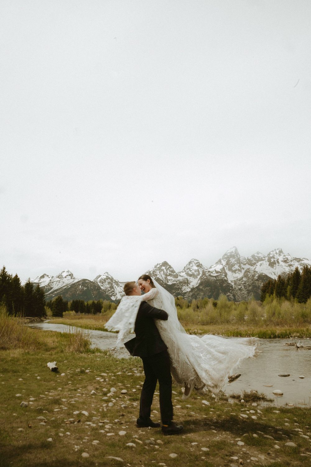 Groom lifts bride in wedding attire with Grand Teton mountains and river in background on overcast day.