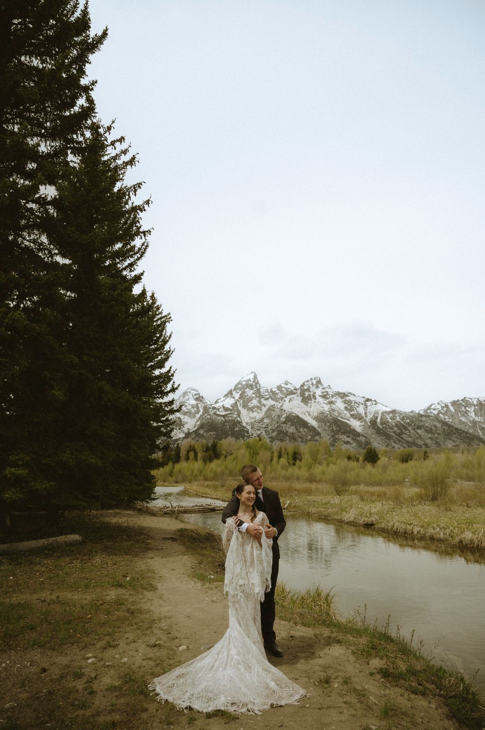 Bride and groom embrace near a mountain lake with snow-capped Grand Tetons and evergreen trees backdrop.