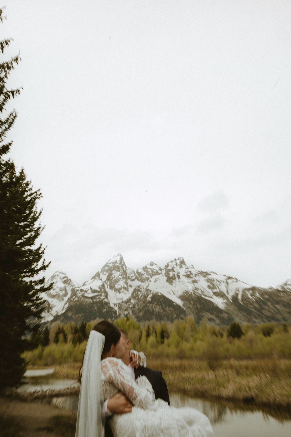 Couple sharing a romantic kiss in lace wedding attire with Grand Teton mountains and forest behind them.