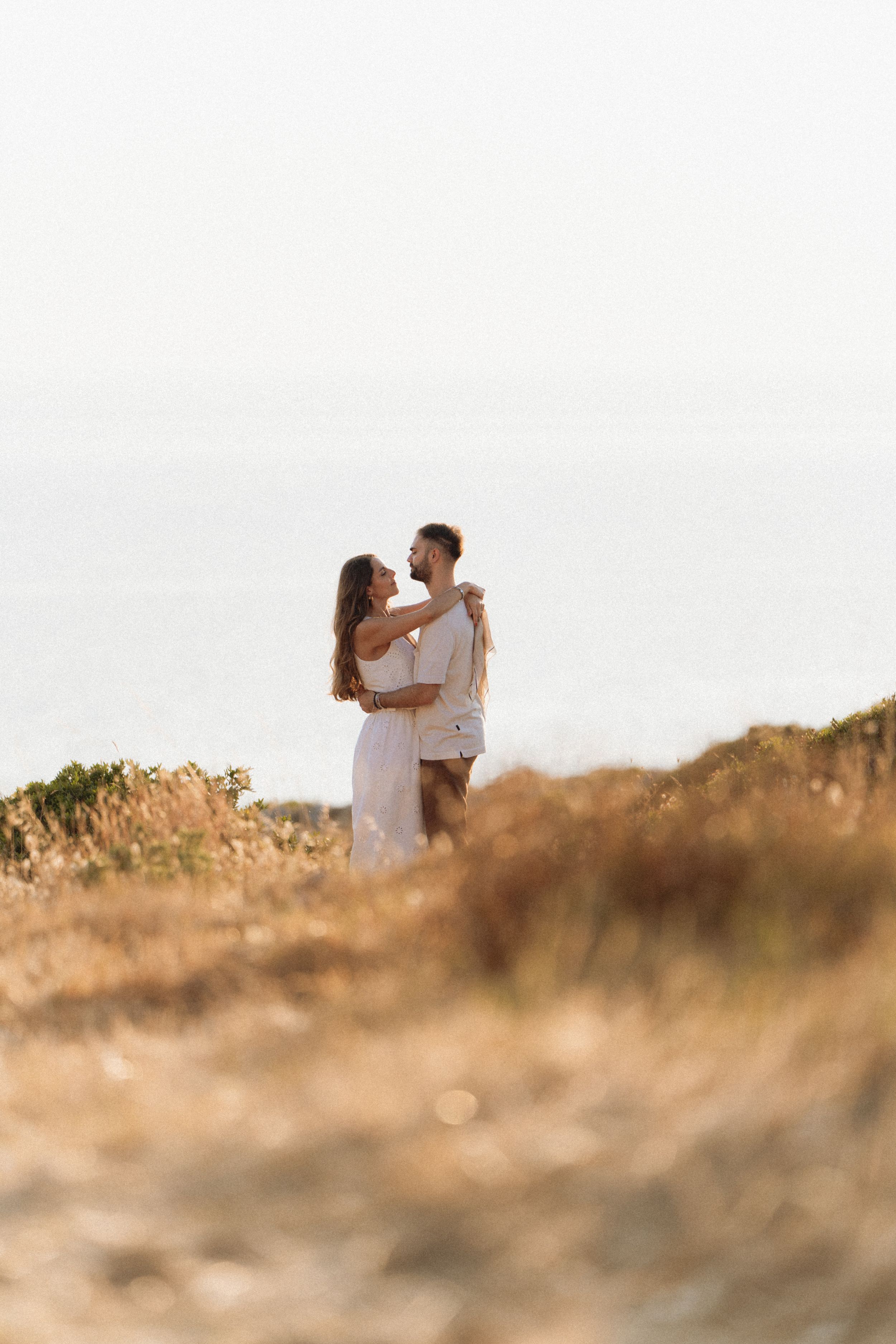 Romantic couple embracing in golden field with ocean backdrop at sunset, woman in white dress.