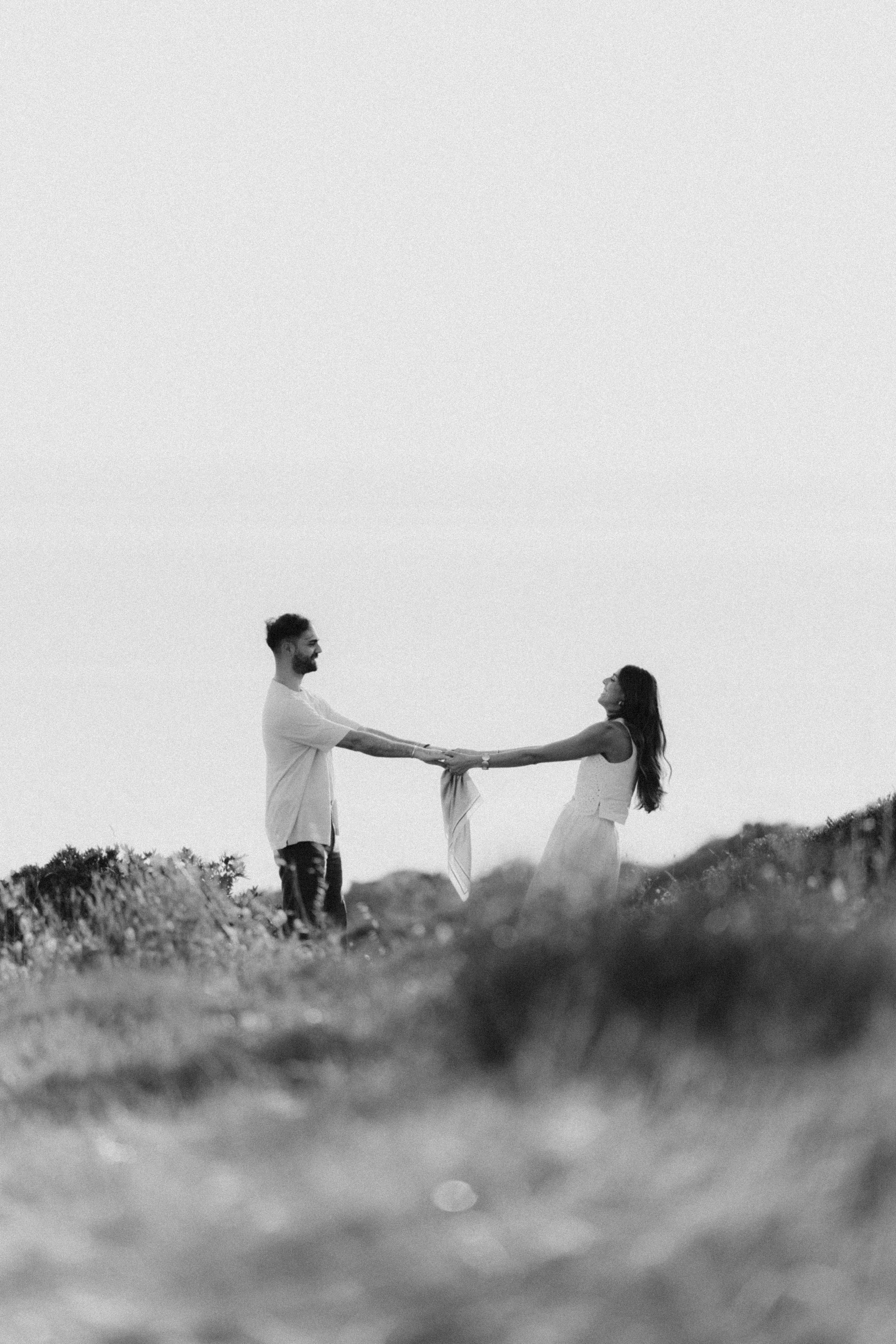 Black and white photo of a couple holding hands in a field, facing each other against a bright sky.