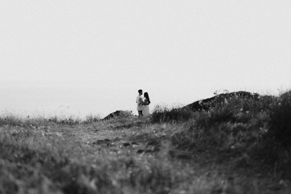 Black and white photo of a couple standing together on a grassy hillside with rocks in a misty landscape.