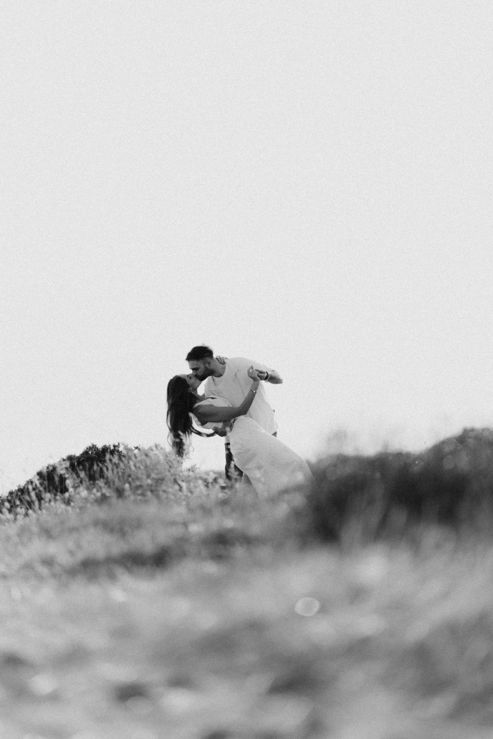 Black and white photo of a couple sharing a romantic kiss on a hilltop in an outdoor field setting.
