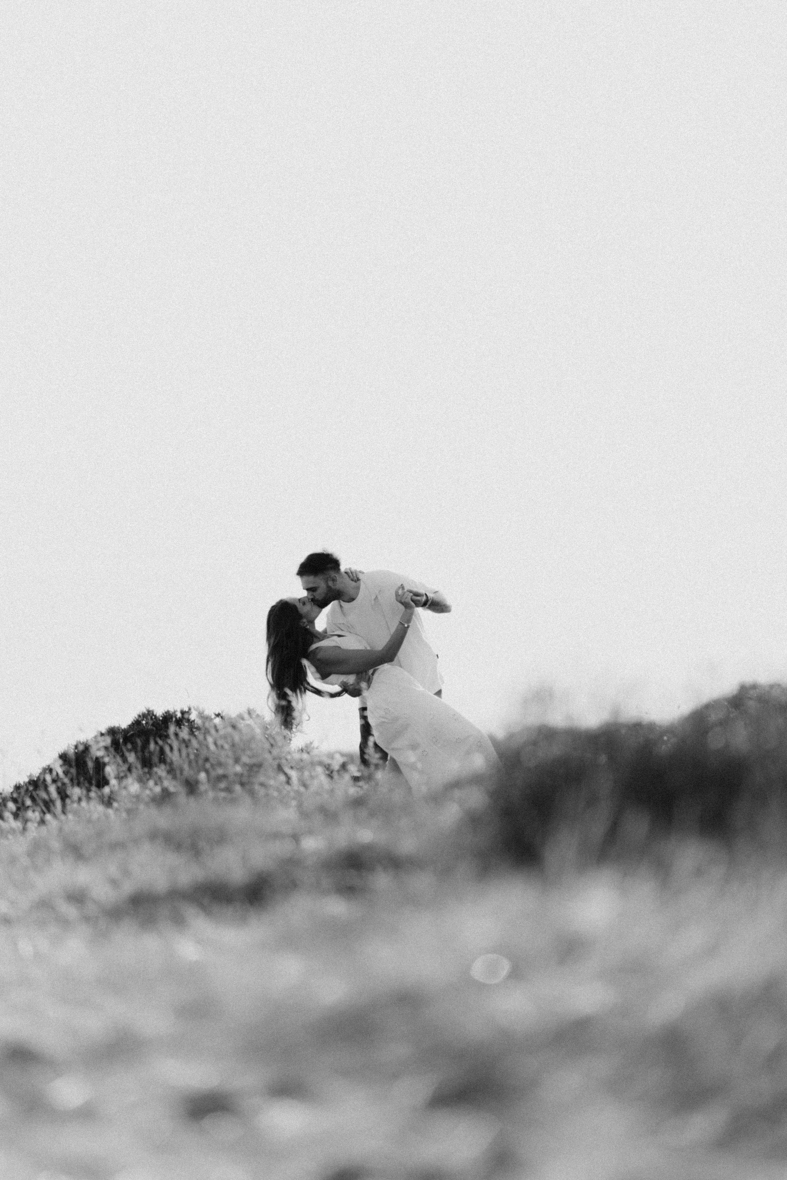 Black and white photo of a couple sharing a romantic kiss on a hilltop in an outdoor field setting.