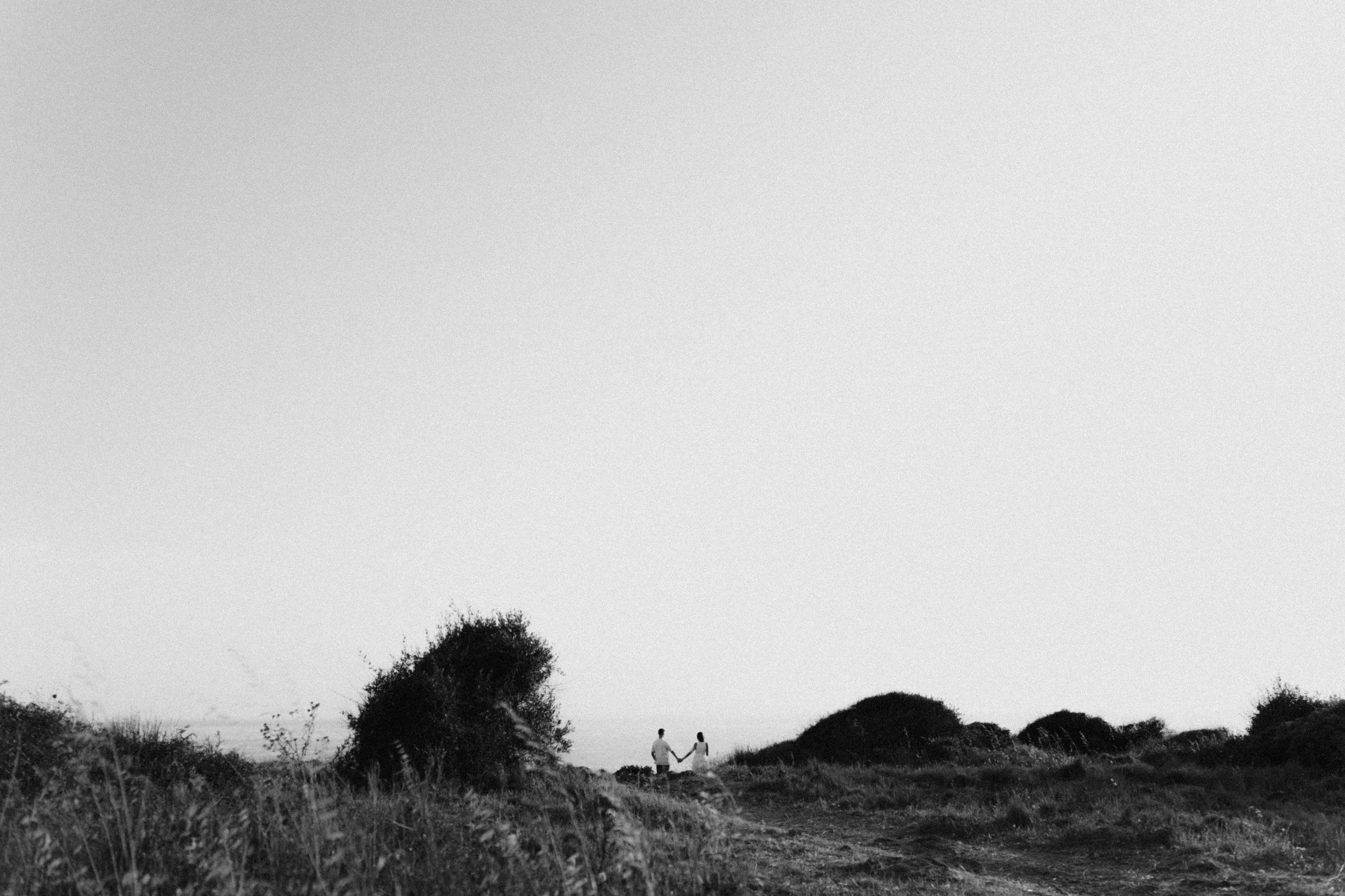 Couple walks through scenic rural landscape with haystacks in minimalist black and white photography.
