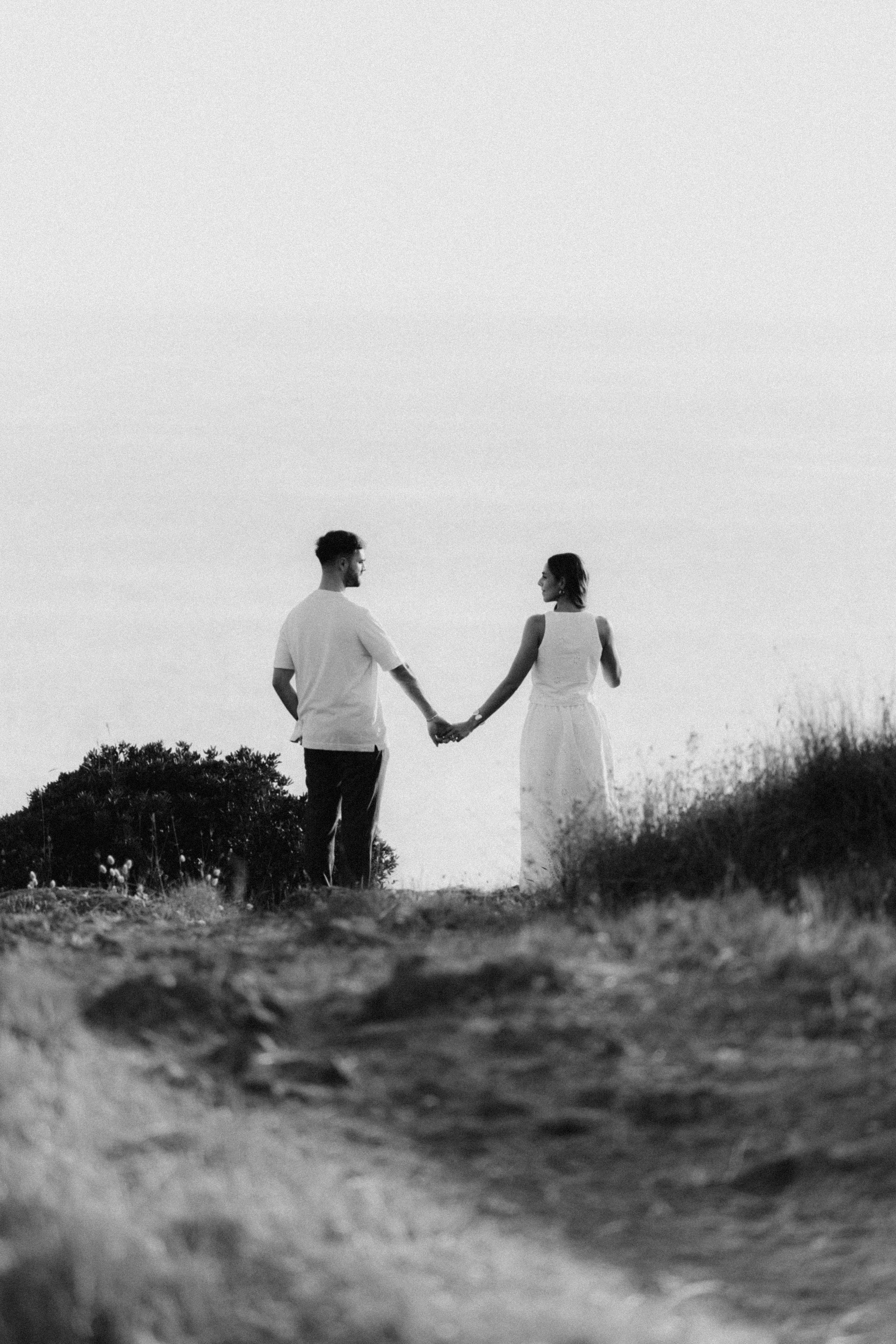 Black and white photo of a couple holding hands on a coastal cliff, gazing at the ocean horizon.