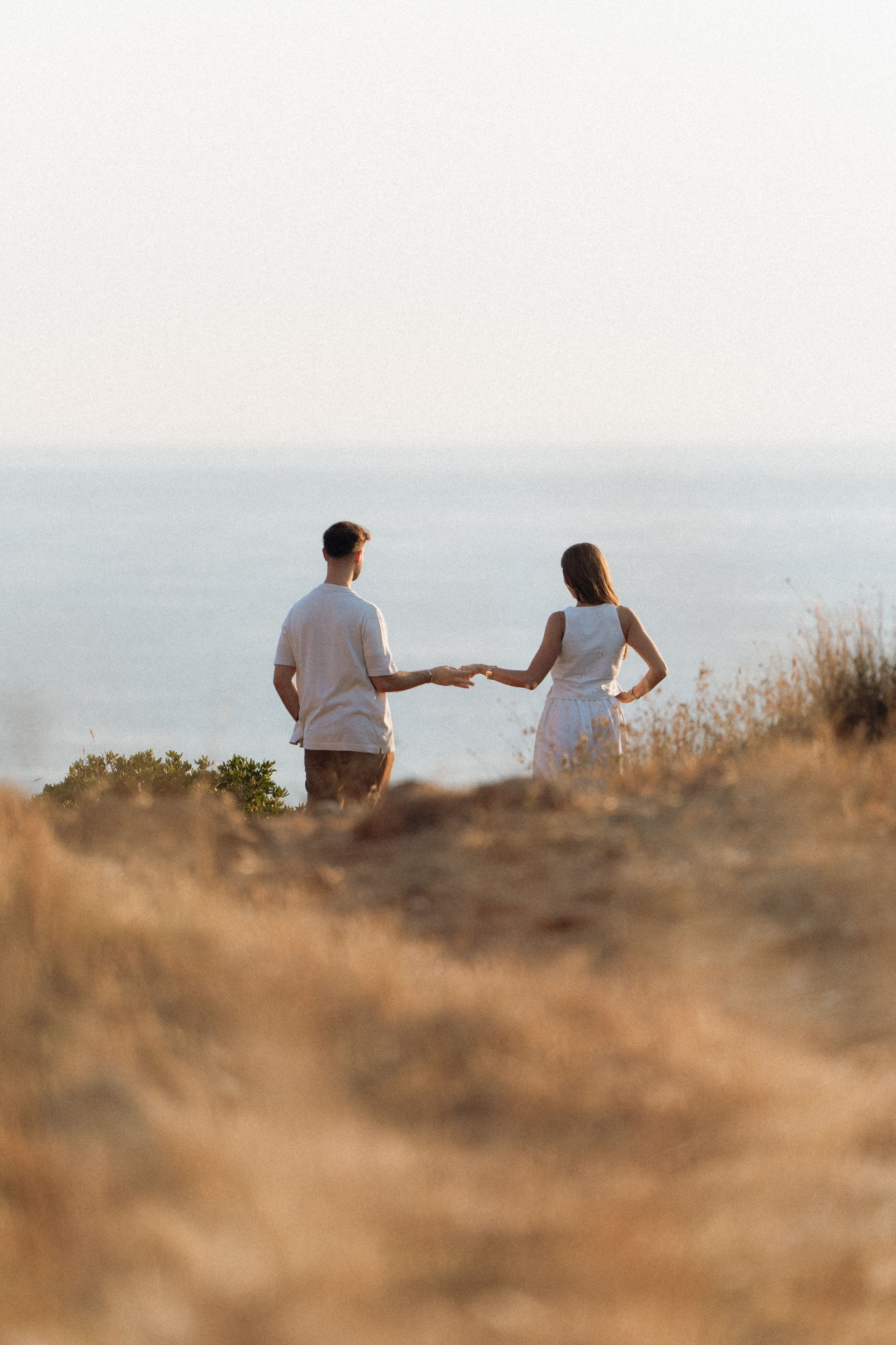 Couple holding hands walking toward ocean view on golden coastal hillside at sunset.