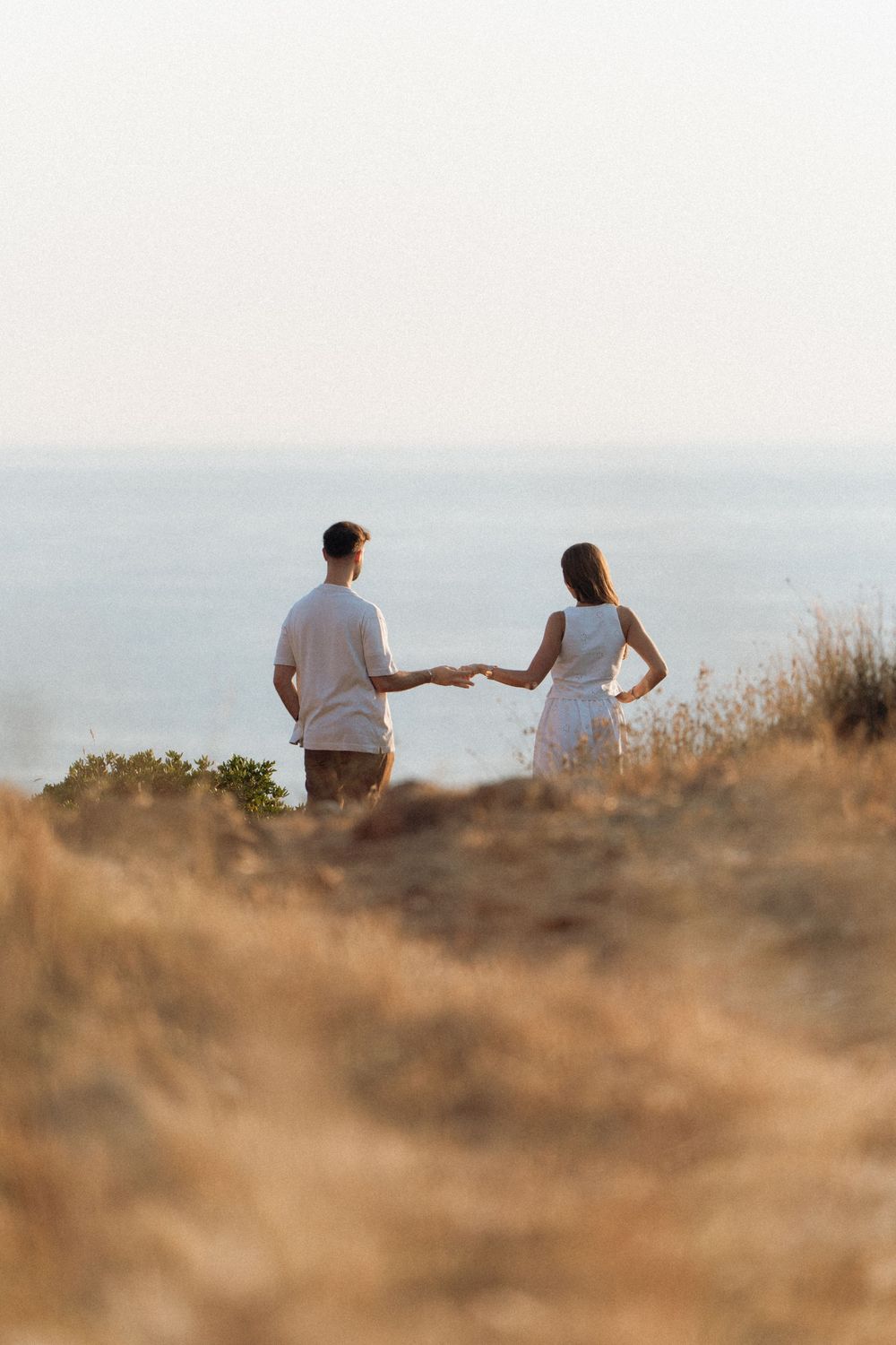 Couple holding hands walking toward ocean view on golden coastal hillside at sunset.