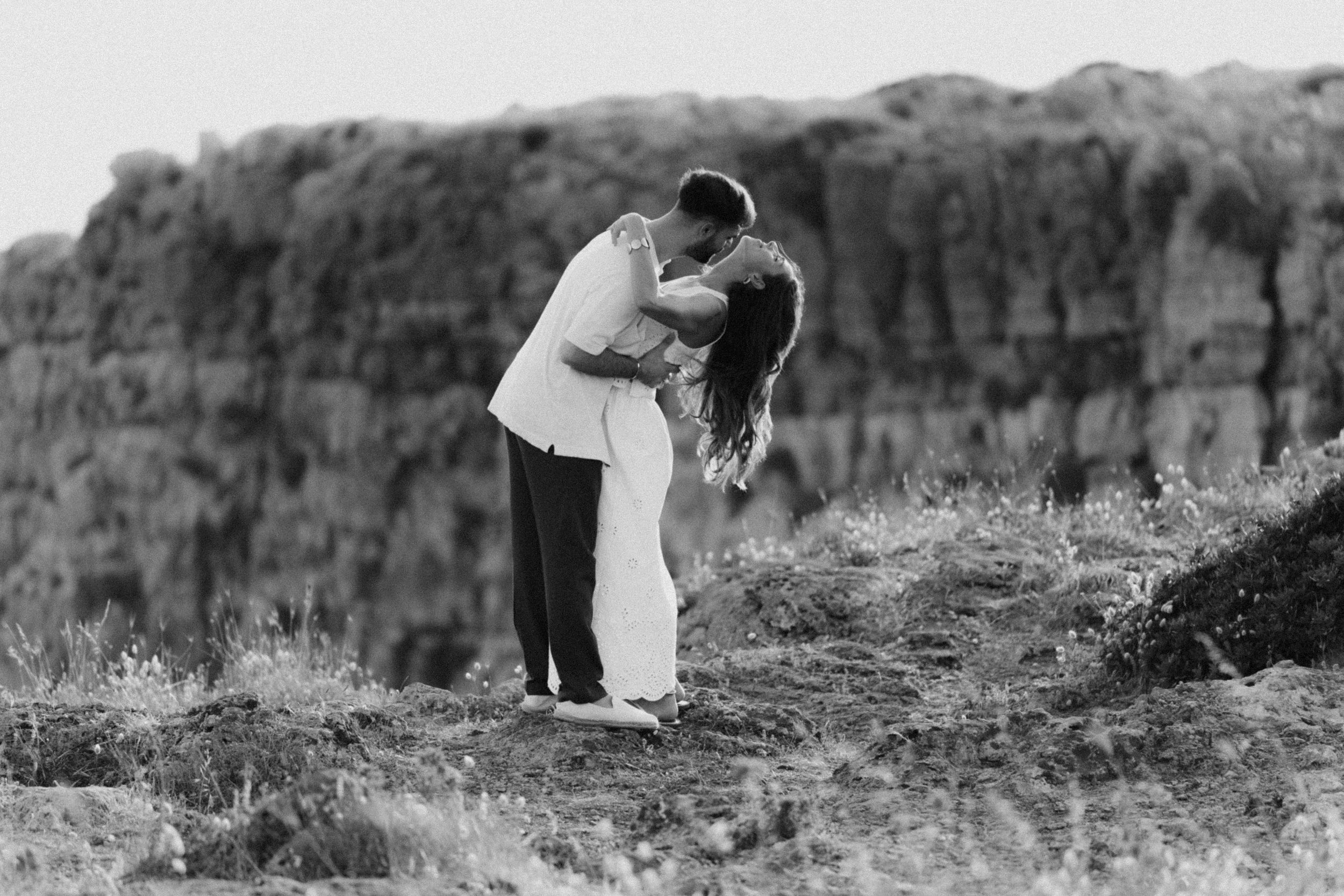 Black and white photo of a couple kissing romantically in a field with hay bales in the background.