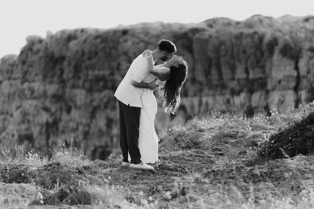 Black and white photo of a couple kissing romantically in a field with hay bales in the background.