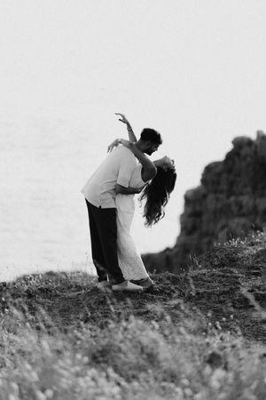 Couple sharing a romantic dip kiss on a coastal cliff in black and white.